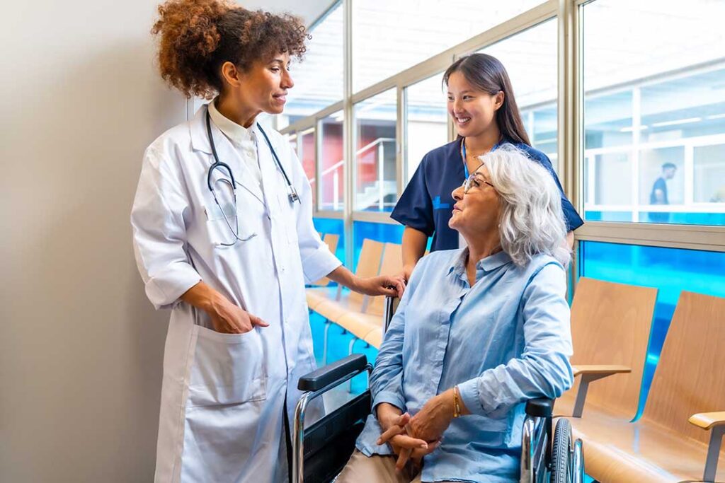 An older adult smiles while chatting with a caregiver at a dining table in a comfortable shared living area.